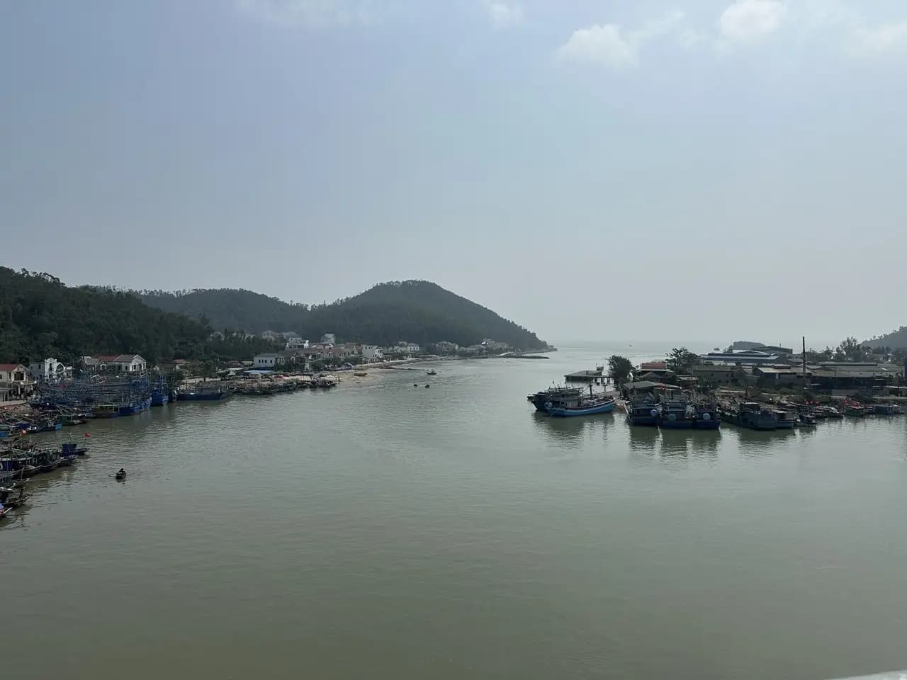 Cửa lạch ở vùng biển Quỳnh nhìn từ trên cầu: thuyền cá sơn xanh neo kín dọc hai bên bờ, sau lưng là xóm nhà mái đỏ dưới chân đồi cây, phía xa lòng lạch mở ra biển