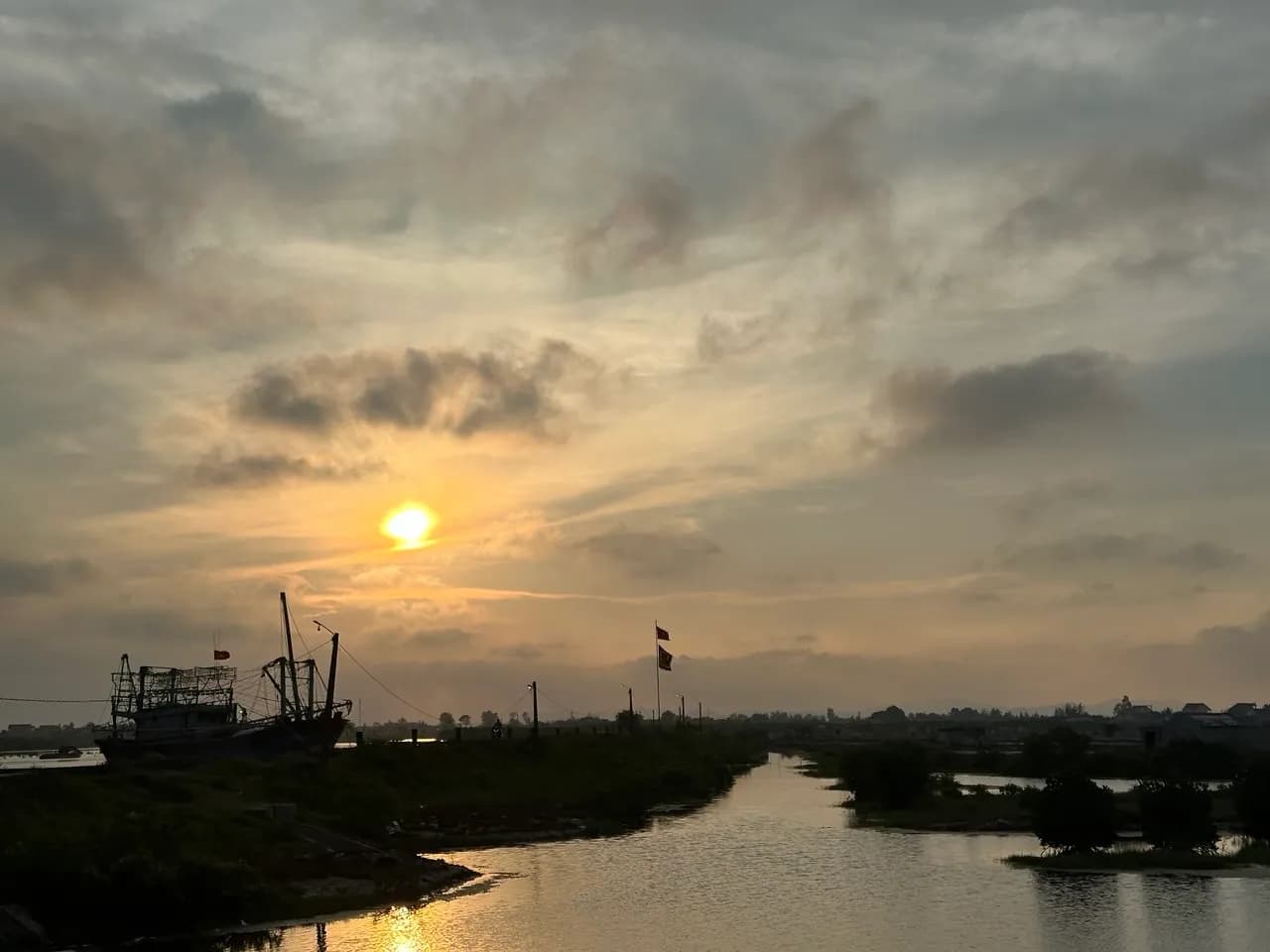 Hoàng hôn trên lạch gần bãi Quỳnh Nghĩa, một thuyền cá gỗ neo bên bờ, mặt trời phản chiếu xuống mặt nước